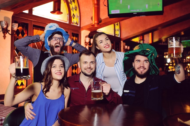 Group or company of friends - young guys and girls holding glasses of beer, watching football, laughing and smiling at the bar during the Oktoberfest festival