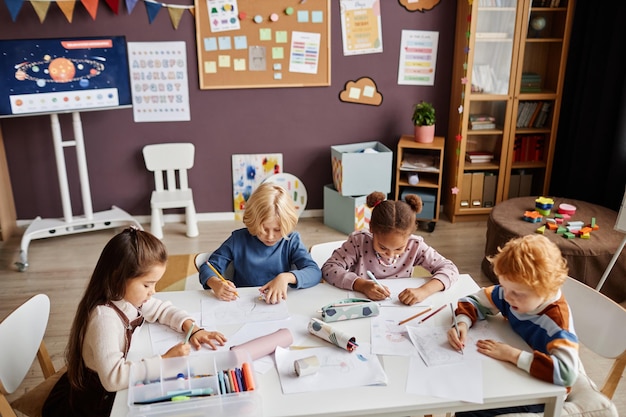 Group of creative little learners of nursery school gathered by table drawing with multicolor crayon