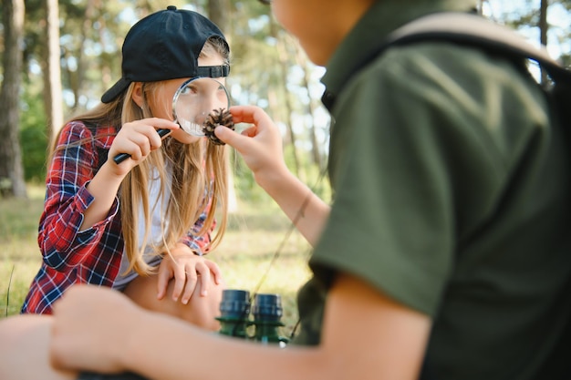 Gruppo di curiosi ragazzi della scuola felice in abiti casual con zaini che esplorano la natura e la foresta insieme nella soleggiata giornata autunnale ragazza che tiene la lente d'ingrandimento e guardando il cono di abete nelle mani del ragazzo