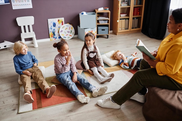 Group of curious intercultural kids relaxing on carpet in front of teacher reading them fairy tale i