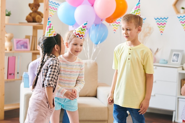 Group of cute little kids in birthday caps discussing rules of new game while having fun at home party in decorated room