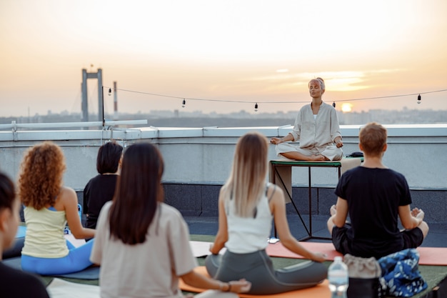 Group of different people are doing meditative yoga practices at the rooftop on a beautiful sunset at summer evening