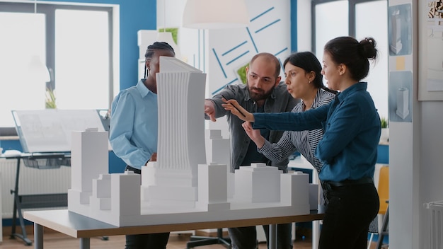 Group of diverse colleagues pointing at building model to work on architectural urban project. Team of people working on maquette design to plan construction layout for development.