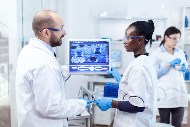 Group of diverse healthcare experts doing virus examining on computer. Multiethnic team of medical researchers working together in sterile lab wearing protection glasses and gloves.