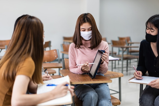 Group of diverse international students wearing protective  masks and talking, discussing project, sitting at desk in the classroom at the university