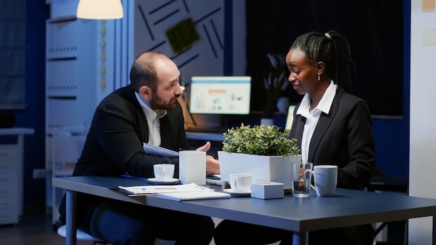 Photo group of diverse managers businesspeople analyzing company graphs paperwork brainstorming company ideas in office meeting room late at night