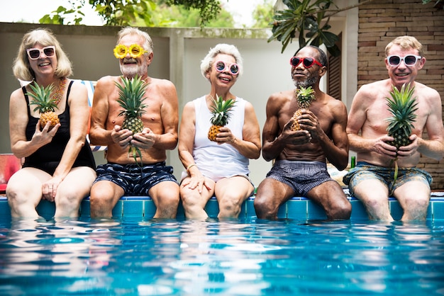 Photo group of diverse senior adults sitting at poolside holding pineapples together