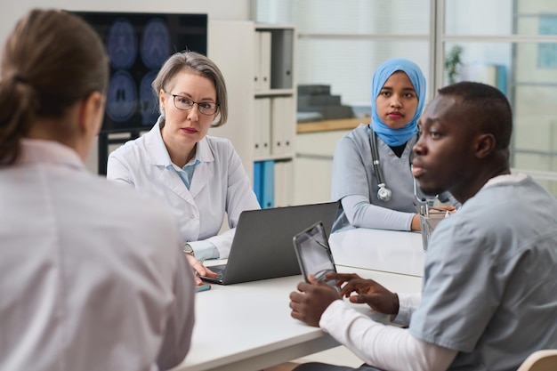 Group of doctors listening to their colleague while using gadgets during teamwork at meeting