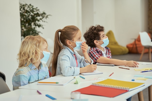 Group of elementary school kids wearing protective mask during coronavirus pandemic listening to