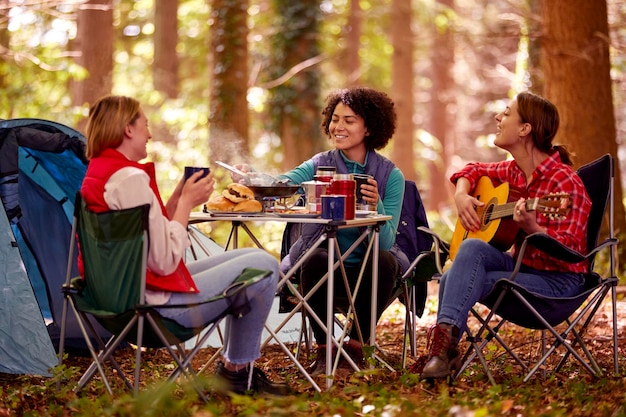 森でキャンプ休暇中の女性の友人のグループは、食事を食べ、ギターと一緒に歌う