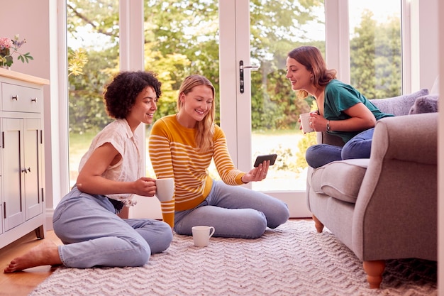 Photo group of female friends relaxing at on sofa at home looking at mobile phone and drinking coffee