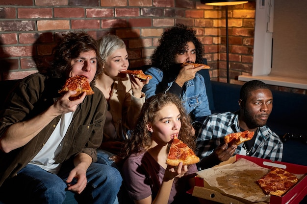 Группа из пяти молодых друзей сидит на диване у себя дома, ест пиццу, смотрит телевизор, интересный комедийный фильм. концепция дружбы, еды и отдыха