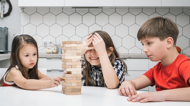 Group of focused children playing board game Jenga building tower made of wooden blocks beams Worried girl cover eyes