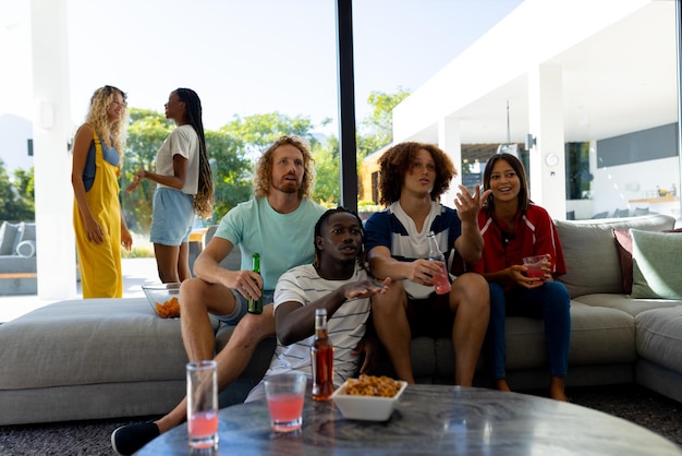 Group of focused diverse friends sitting on sofa, watching sport on tv and drinking