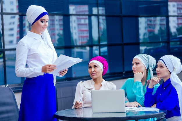 Group of four beautiful and successful Muslim business rich women perfect fashion look makeup and turban meeting in a summer restaurant on street a female person gives a presentation to colleagues