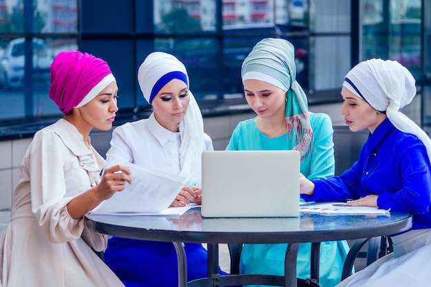 Group of four caucasian Muslim, office lady discussing with a businesswoman partner job interview shawl and turban on head perfect skin and makeup sitting at a round table with a laptop and papers.