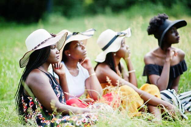 Group of four gorgeous african american womans wear summer hat sitting at green grass in park.