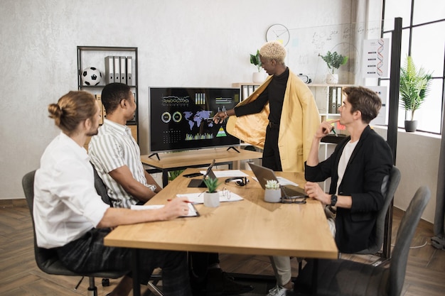 Group of four partners sitting at desk and listening speech of african american man near monitor