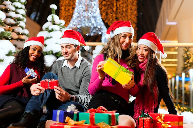 Group of four people with Santa hats sitting amid artificial snow covered fir trees and lights 