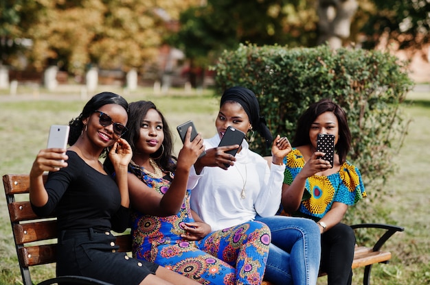 Group of four women sitting on bench outdoor with mobile phones at hands and making selfie