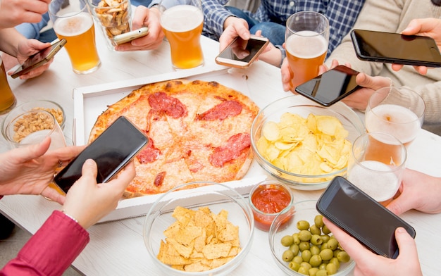 Group of friends are eating pizza, drinking beer and using smartphone in a pub