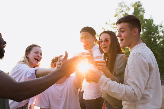 Group of friends clinking beer glasses during picnic at the beach. Lifestyle, friendship, having fun, weekend and resting concept.