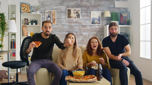 Group of friends happy after their favorite football team wins the championship. Friends sitting on sofa eating pizza and chips.