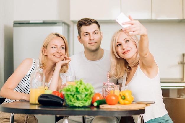 Group of friends having fun in kitchen, smiling taking selfie, preparing vegetable salad. Selective focus. Social media, technology, cooking, students concept.