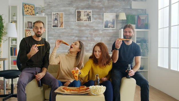 Group of friends laughing hard while a sitcom on tv in living room. Pizza and popcorn on table.