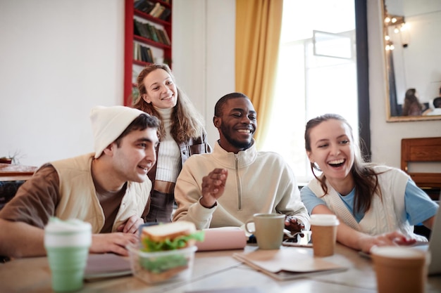 Group of friends laughing and looking something on laptop while sitting at the table in the room