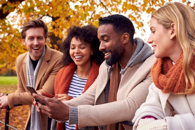 Photo group of friends outdoors wearing coats and scarves looking at photos on phone in autumn park