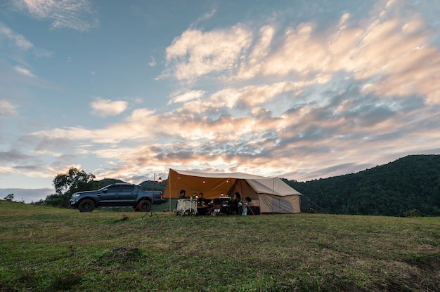 Group of friends relaxing inside large tent and pickup parked on hill in countryside at camping holiday