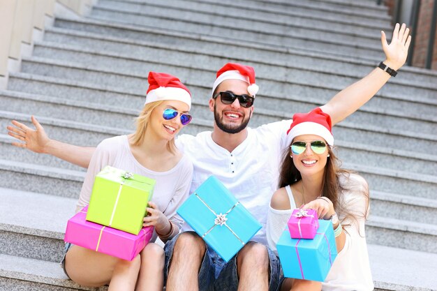 group of friends in Santa's hats sitting in the city with presents