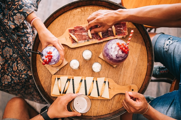 A group of friends sits on the terrace in the summer behind a wooden barrel with cocktails and a snack
