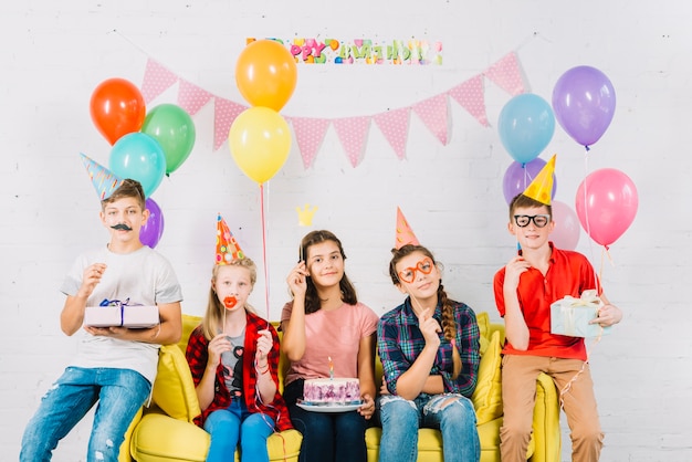 Gruppo di amici seduti sul divano con la torta di compleanno; palloncini e regali