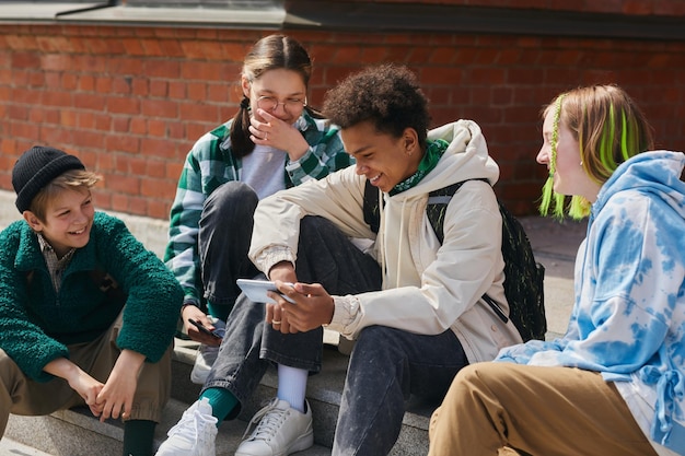 Group of friends sitting on steps of building outdoors and laughing while watching funny video on mo