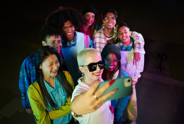 Gruppo di amici che si fanno selfie con il cellulare divertendosi alla festa di notte folla di felici mul...