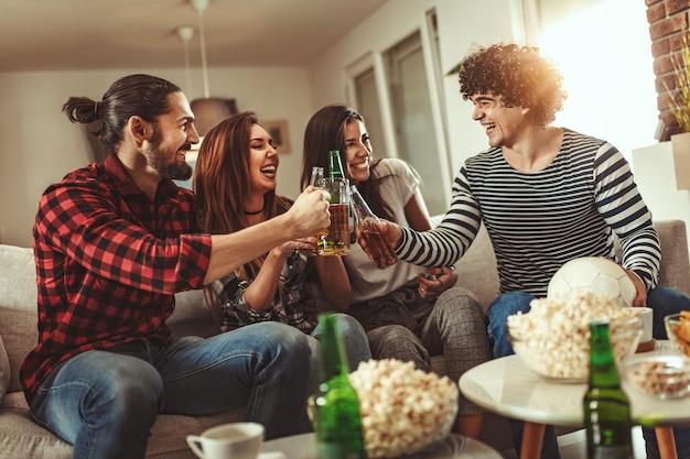 A group of friends watches the broadcast of a sports event. They sit in front of the TV in the living room, eat snack, drink beer, and cheer for the favorite team.