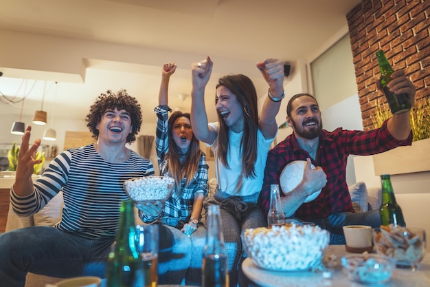 A group of friends watches the broadcast of a sports event. They sit in front of the TV in the living room, eat snack, drink beer, and cheer for the favorite team.