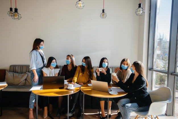 A group of girls in masks sit in a cafe and work on laptops. Teaching students.