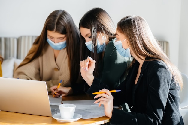 A group of girls in masks sit in a cafe and work on laptops. Teaching students.