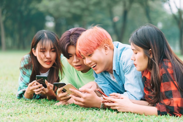 幸せなアジアの友人のグループがスマートフォンを使用して座って、公園でおしゃべりしています