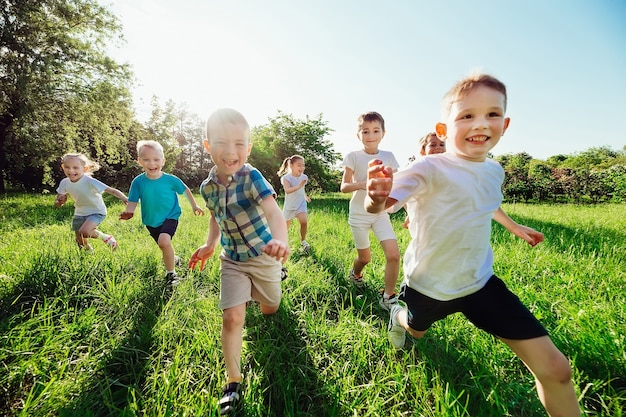 晴れた夏の日に、男の子と女の子の幸せな子供たちのグループが芝生の上の公園を走ります。