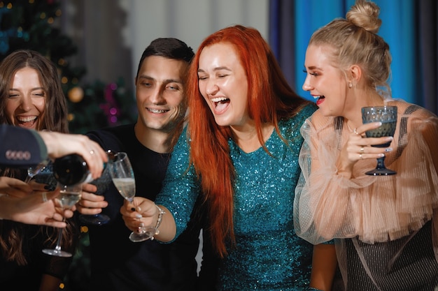 Group of happy friends celebrating Christmas or New Year. A man pours champagne into glasses. Women laugh, sitting at a table near the Christmas tree.