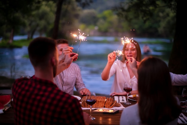 group of happy friends celebrating holiday vacation using sprinklers and drinking red wine while having picnic french dinner party outdoor near the river on beautiful summer evening in nature