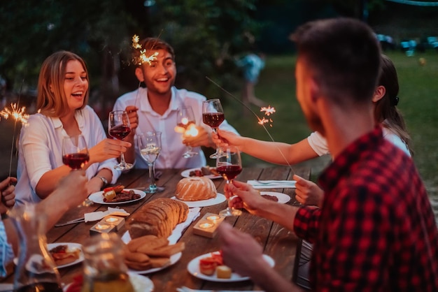 Group of happy friends celebrating holiday vacation using sprinklers and drinking red wine while having picnic french dinner party outdoor near the river on beautiful summer evening in nature.