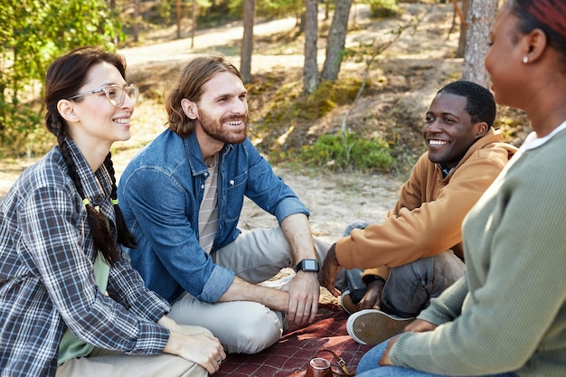 Group of happy friends chatting together sitting on blanket during picnic in the forest