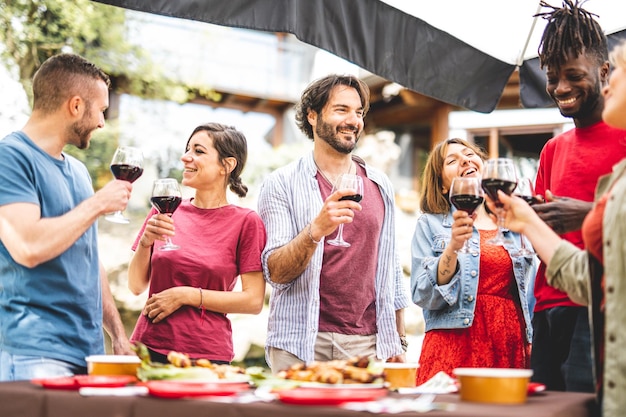 Group of happy friends having fun while drinking red wine and eating food at party focus on young bearded handsome man genuine people