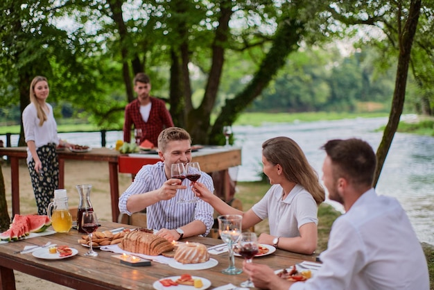 group of happy friends having picnic french dinner party outdoor during summer holiday vacation near the river at beautiful nature