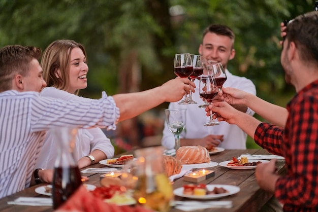 group of happy friends toasting red wine glass while having picnic french dinner party outdoor during summer holiday vacation near the river at beautiful nature